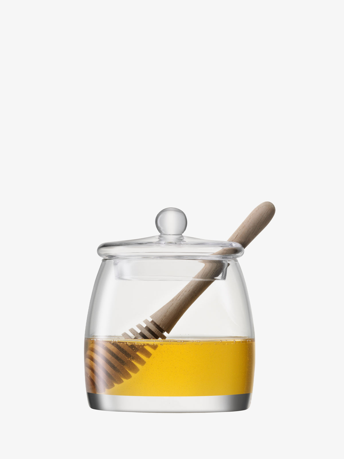 The lsa-international Honey Pot & Dipper features a mouth-blown glass vessel filled with golden honey and finished with a clear glass lid. An oak dipper rests inside, partially immersed in the honey, all set against a plain white background.