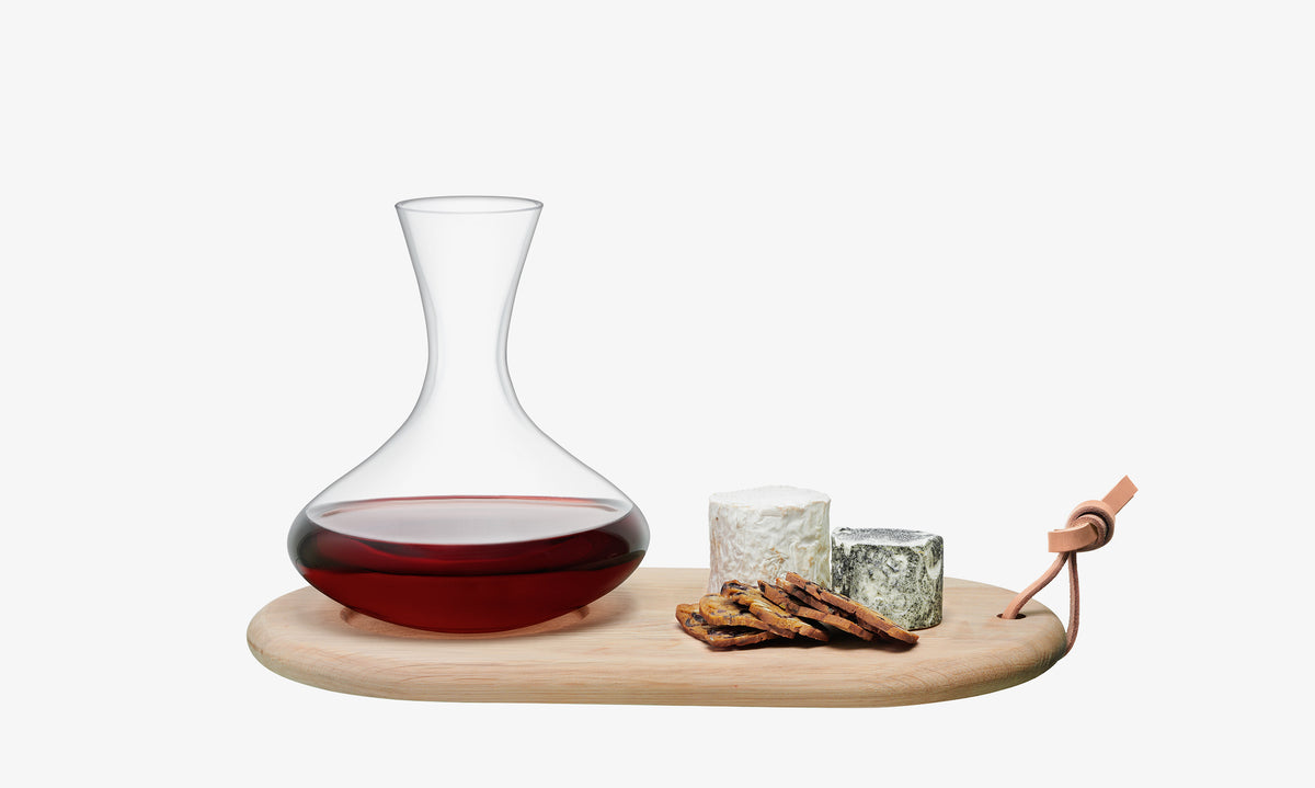 The Carafe & Oak Cheese Board by lsa-international features a glass carafe filled with red wine, accompanied by two varieties of cheese and crackers, all beautifully displayed on an oak board with a leather strap against a white background—perfect for elegant wine aeration and tasting experiences.