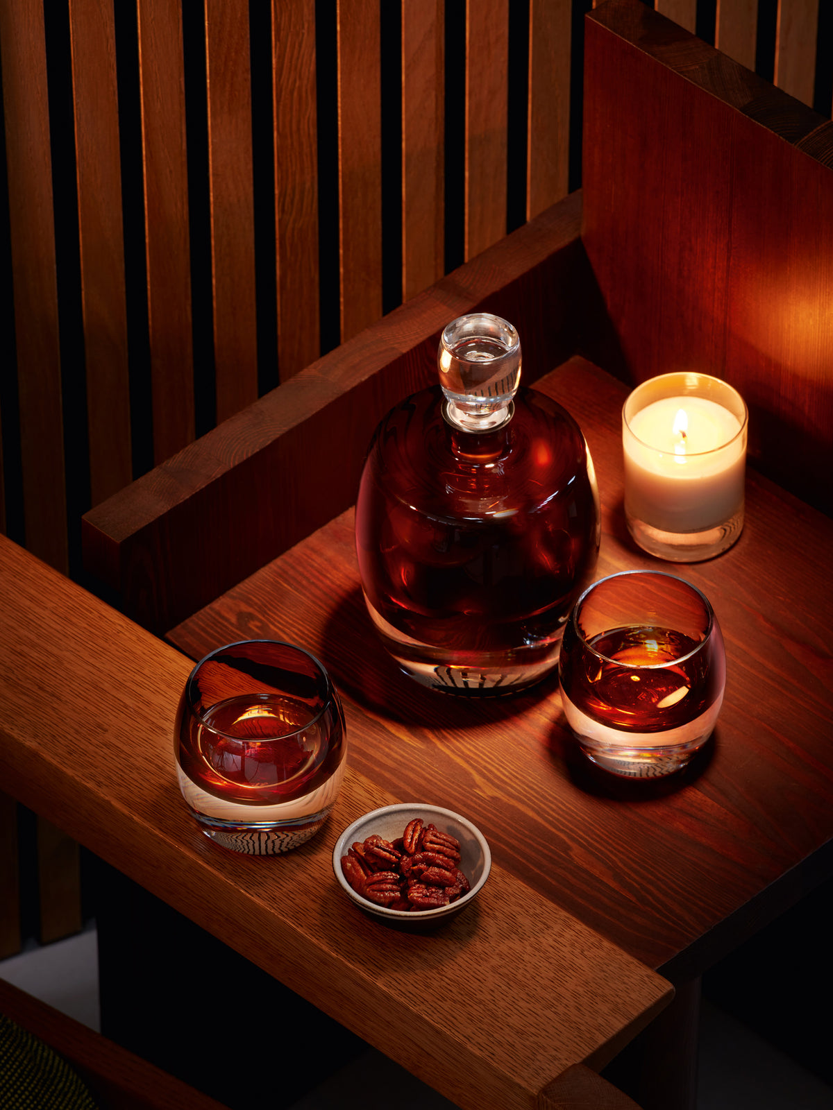 A lsa-international Whisky Set, featuring a decanter and two handcrafted tumblers filled with dark liquor, sits on a wooden table beside a lit candle and a small bowl of pecans, creating a cozy atmosphere.