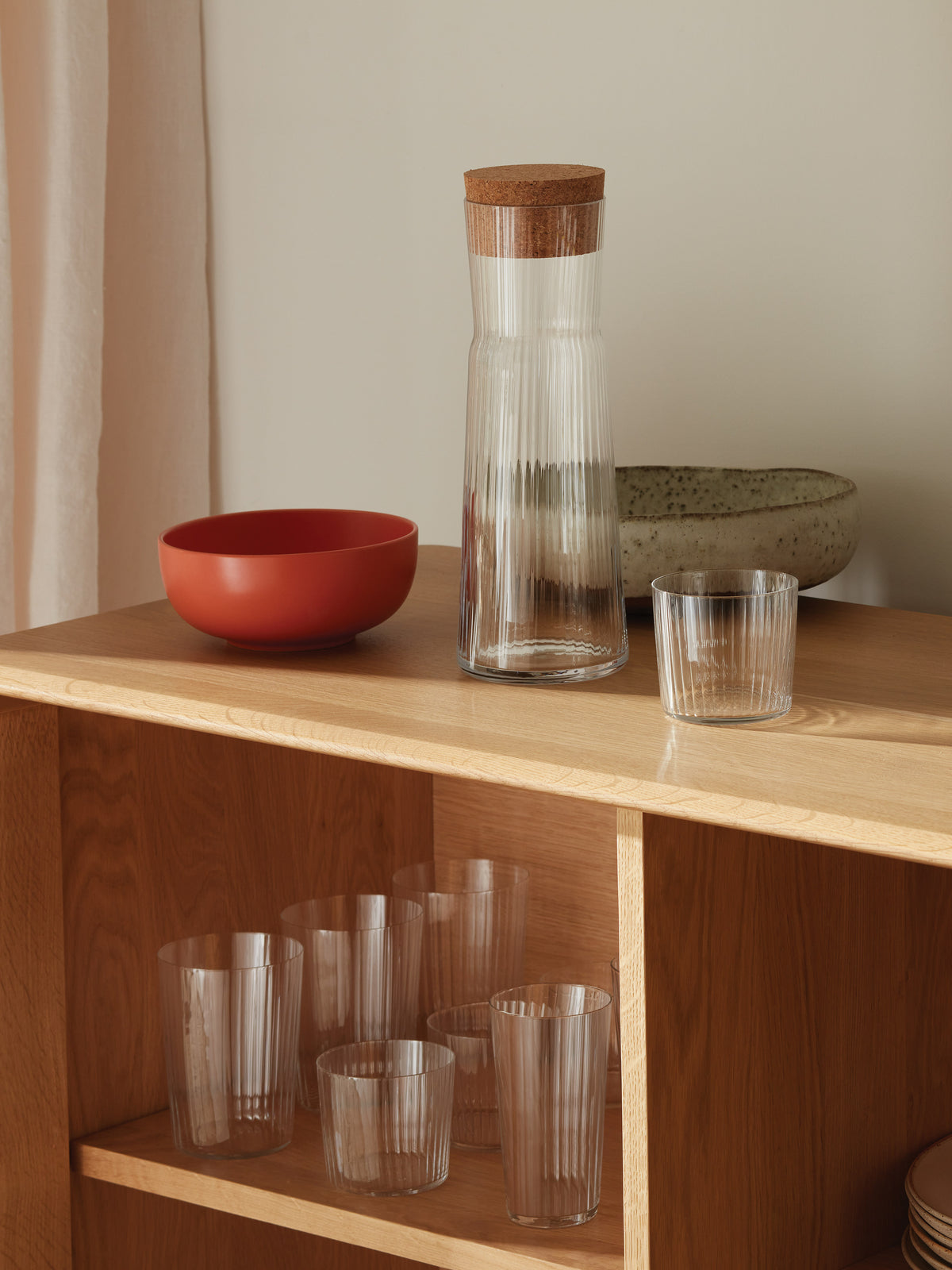 A wooden shelf holds the lsa-international Carafe & Cork Stopper, featuring a fluted clear glass design paired with a matching glass, along with a red bowl and a speckled oval dish. Additional clear glasses are displayed on the lower shelf.
