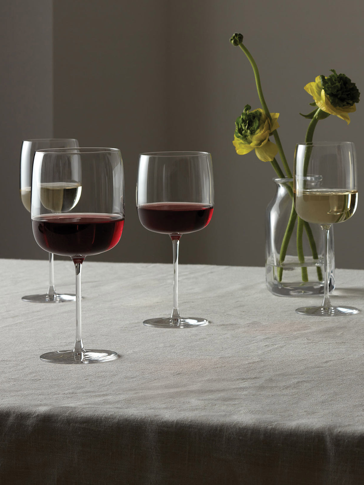 Four lsa-international Wine Glasses with tall stems—two filled with red wine, two with white—rest on a linen-covered table. Crafted from crystalline glass, these elegant pieces reflect the soft lighting beautifully. In the background, a glass vase holding yellow flowers completes the calm and refined atmosphere.