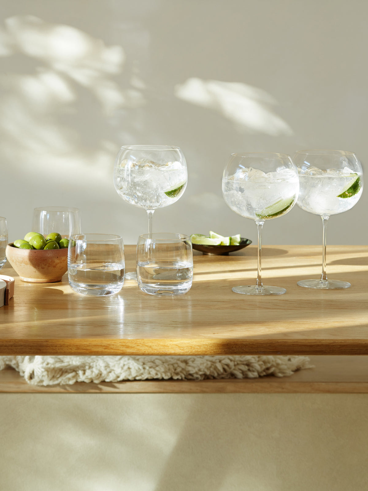 Four lsa-international Balloon Glasses, filled with gin and tonic, ice, and lime, are arranged next to two empty crystalline tumblers. Nearby, a bowl of green olives and a plate of lime wedges sit on a wooden table bathed in warm sunlight. Gentle leaf shadows create delicate patterns on the light wall behind.