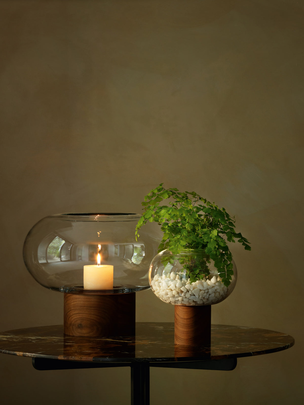 Two mouth-blown LSA International vases on walnut bases: one holding a lit candle and the other containing a small fern planted in white pebbles.