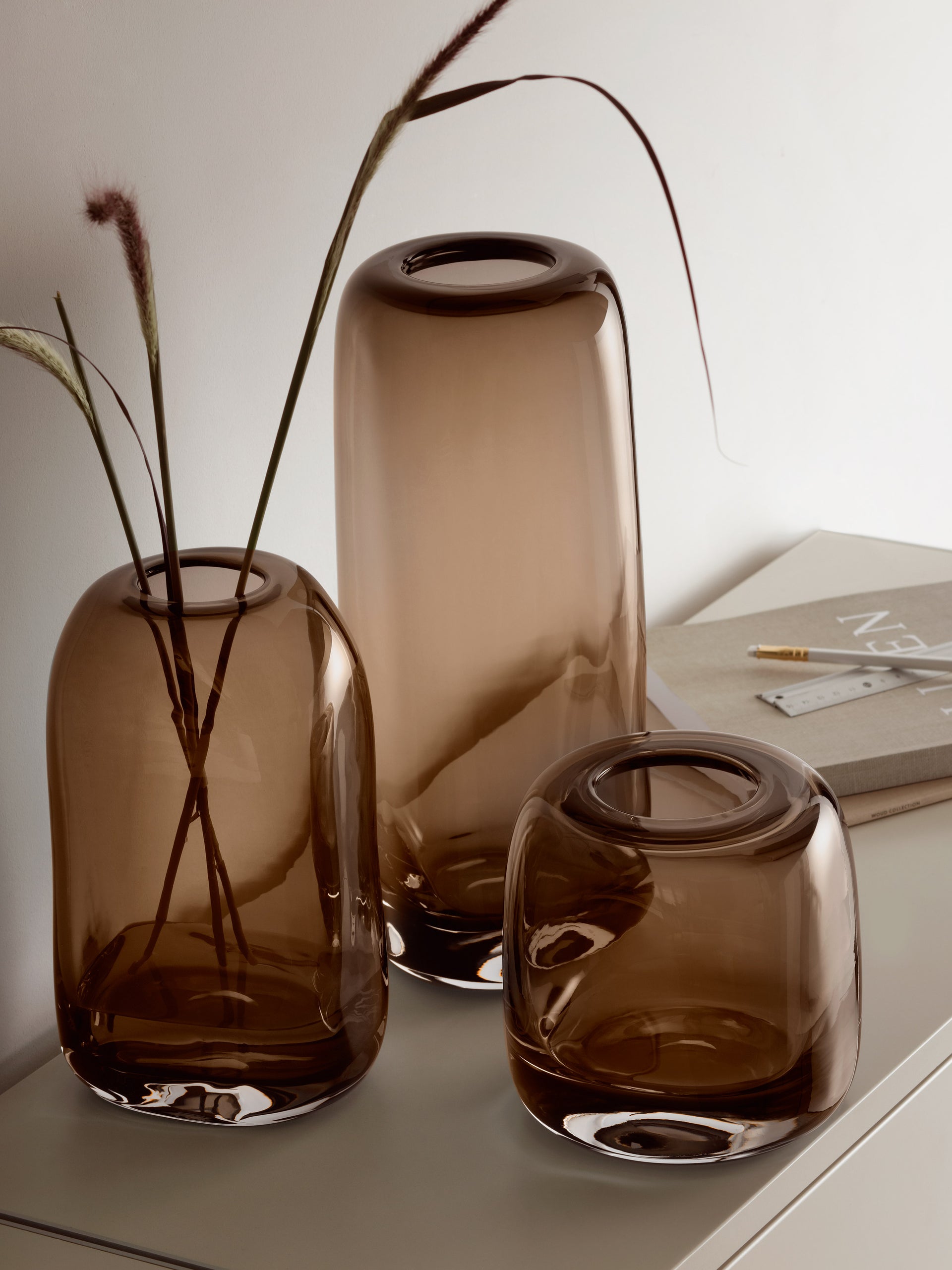 Three contemporary brown lsa-international Vases, crafted from mouth blown glass in varying heights, rest on a light surface. The tallest Vase displays a few dried, wispy grass stems. Beside the Vases lie two closed books and two gold paperclips.