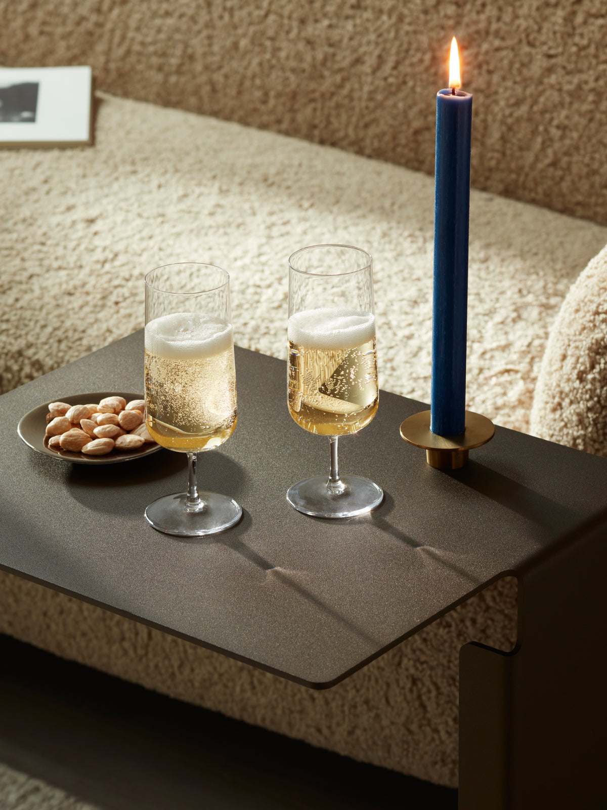 Two lsa-international Champagne Flute glasses filled with sparkling wine, a plate of almonds, and a lit blue candle rest on a dark table in front of a textured beige sofa.