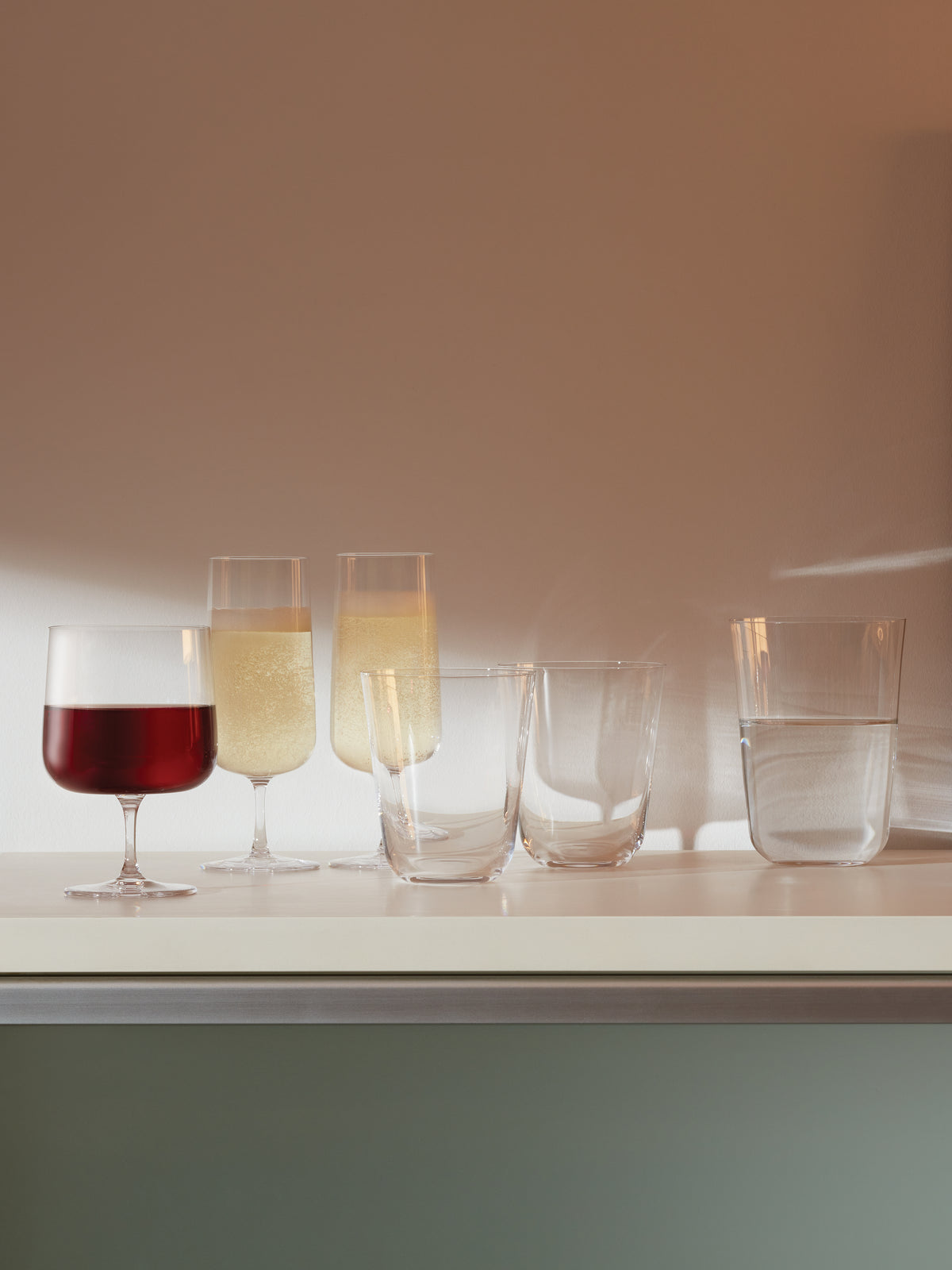 Five lsa-international Wine Glasses with short stems are arranged on a white surface: one filled with red wine, two with sparkling wine, one empty, and one with water. Crafted from lead-free crystalline glass, these Wine Glasses catch the soft light and cast subtle shadows on the wall behind them.