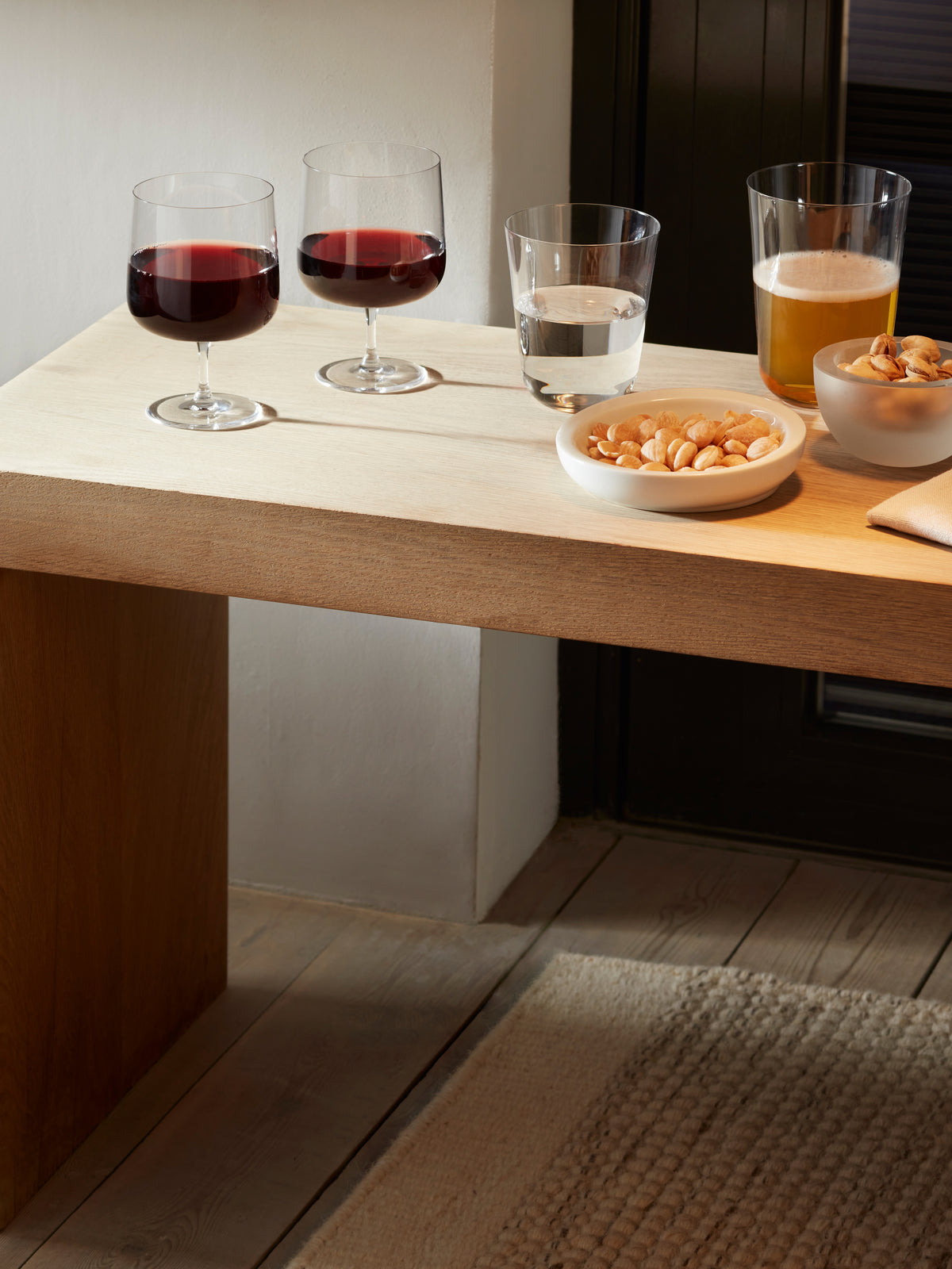 A wooden table near a window displays two lsa-international Wine Glasses filled with red wine, alongside a glass of water, a glass of beer, and two bowls of almonds.