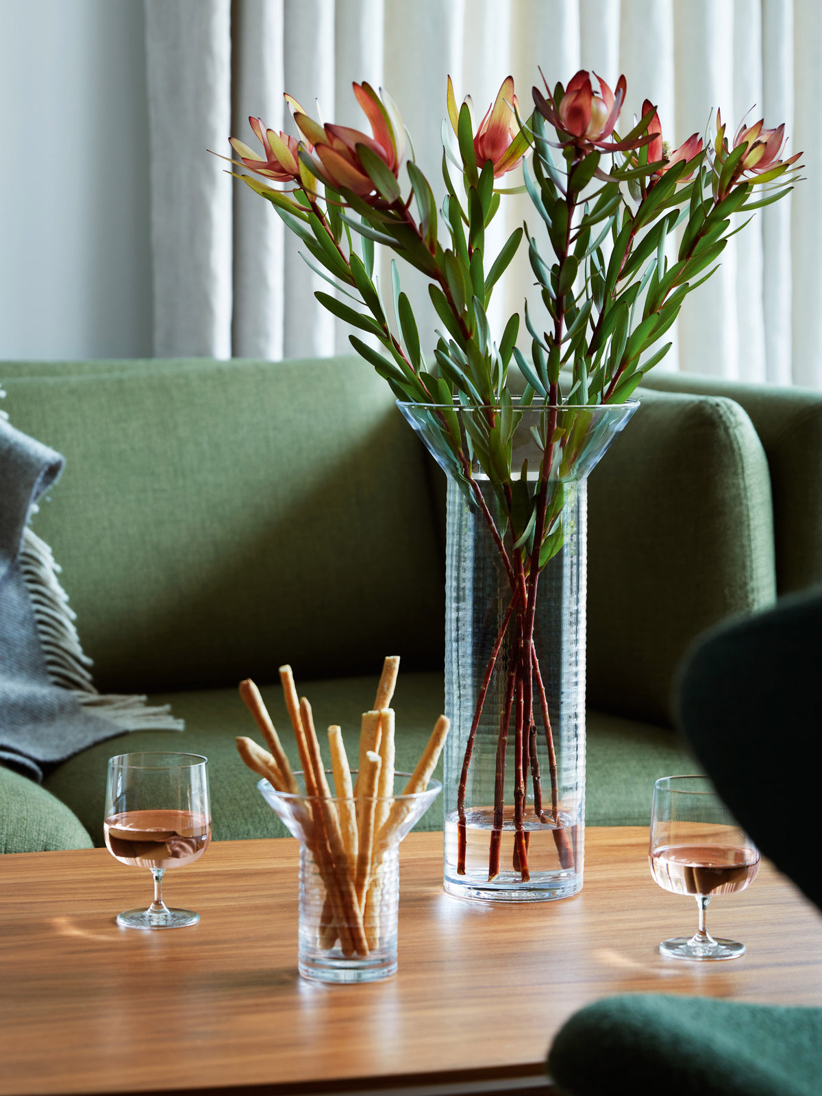 A tall, mouth-blown contemporary lsa-international vase holds stems of protea flowers on a wooden coffee table, flanked by two glasses of rose wine and a small bowl of breadsticks.
