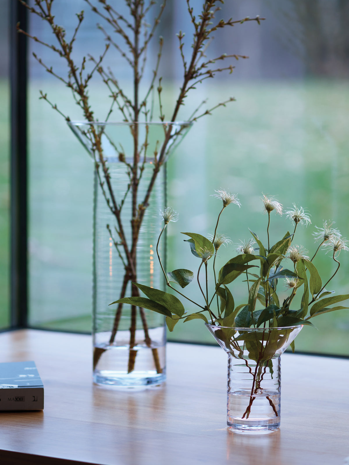 Two mouth-blown, contemporary LSA International vases of different heights sit on a wooden surface, each holding small branches or leafy stems.