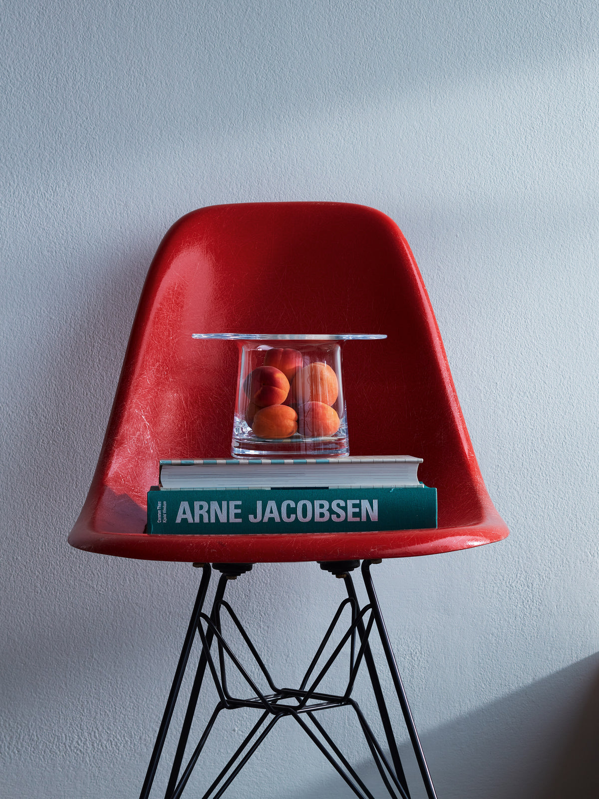 A mouth-blown lsa-international Vase/Lantern filled with peaches sits on a stack of books on a red molded chair, showcasing contemporary design.