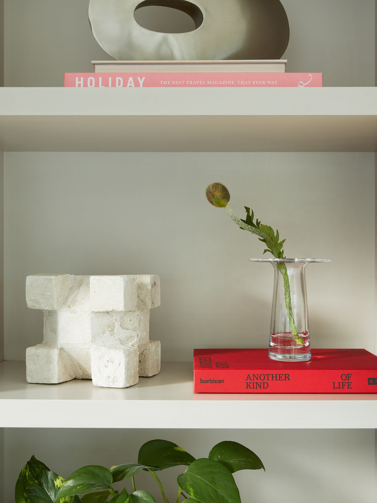 A clear, mouth-blown LSA International Victoria vase holding a single poppy stem, placed on a red book next to a geometric stone sculpture on a bookshelf.
