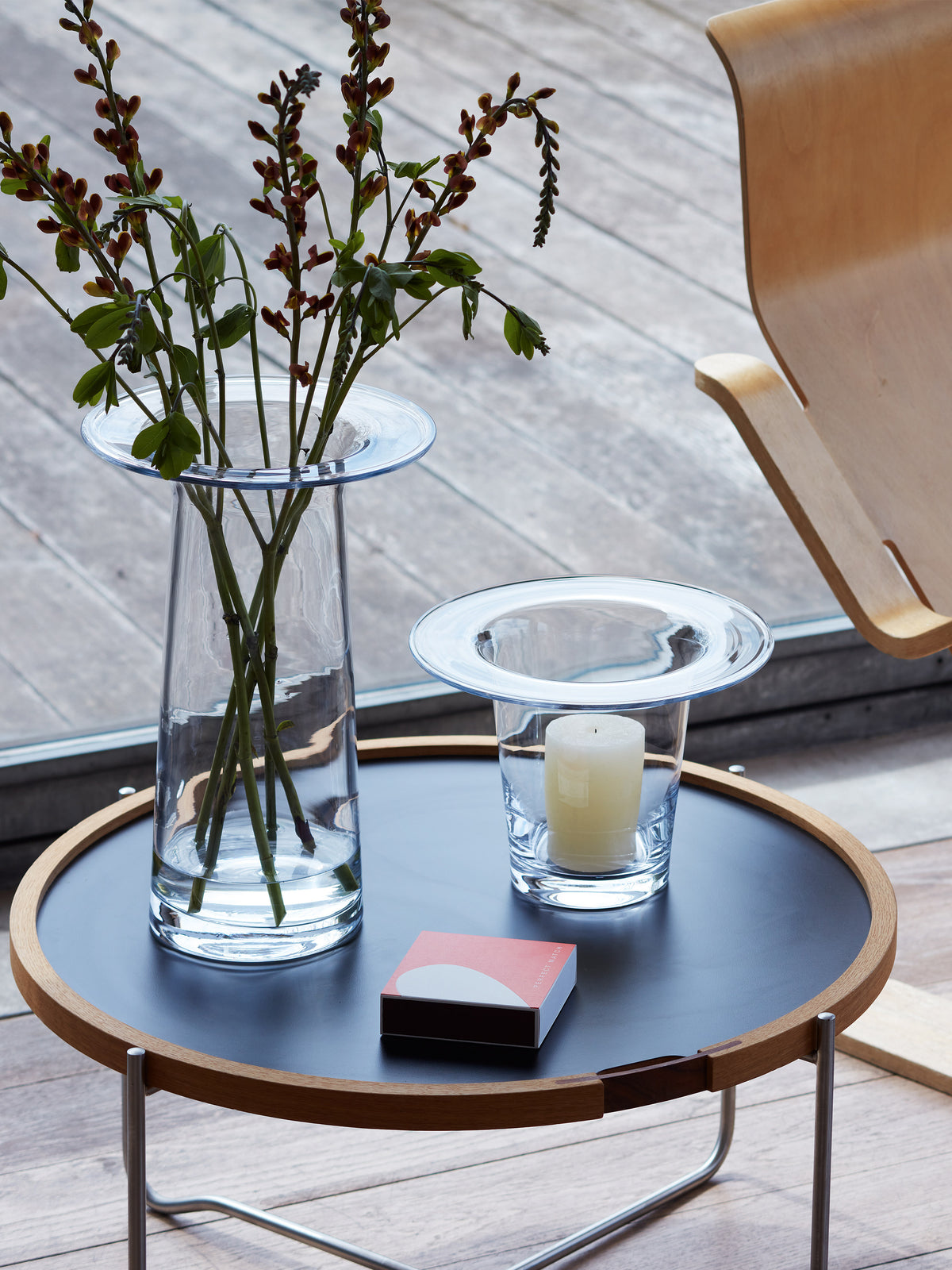 A tall LSA International Vase/Lantern with stems and a shorter glass lantern with a pillar candle sit on a round black table next to a small matchbox.