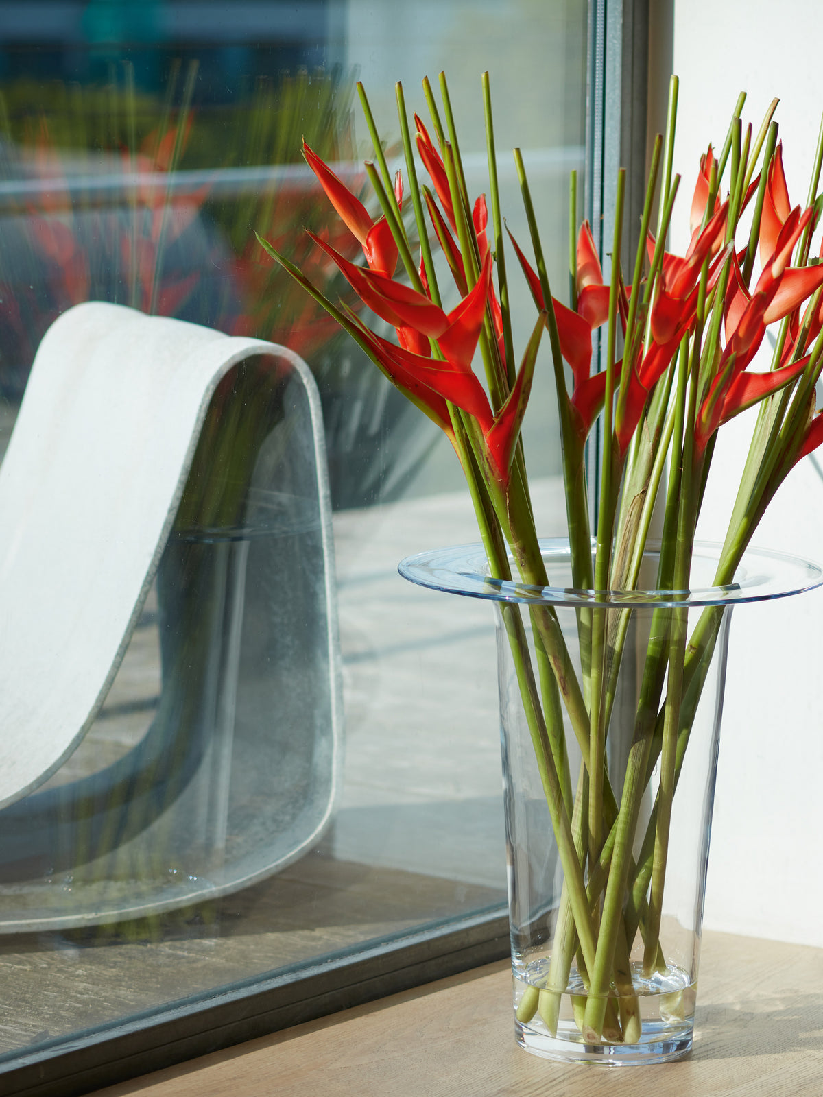 A large, mouth-blown LSA International Vase/Lantern filled with bright red heliconia flowers serves as a statement flower arrangement next to a window.