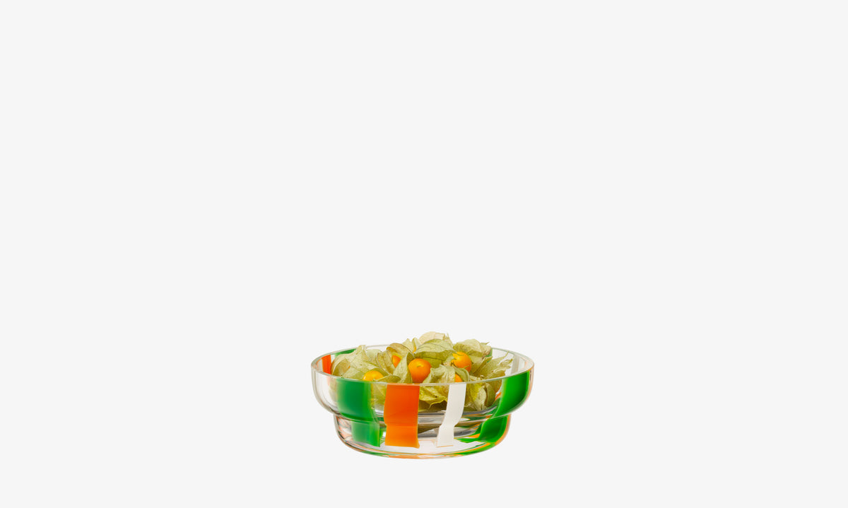 The lsa-international Bowl, a clear mouth-blown piece with orange, green, and white stripes, holds small green fruits in papery husks on a white background.
