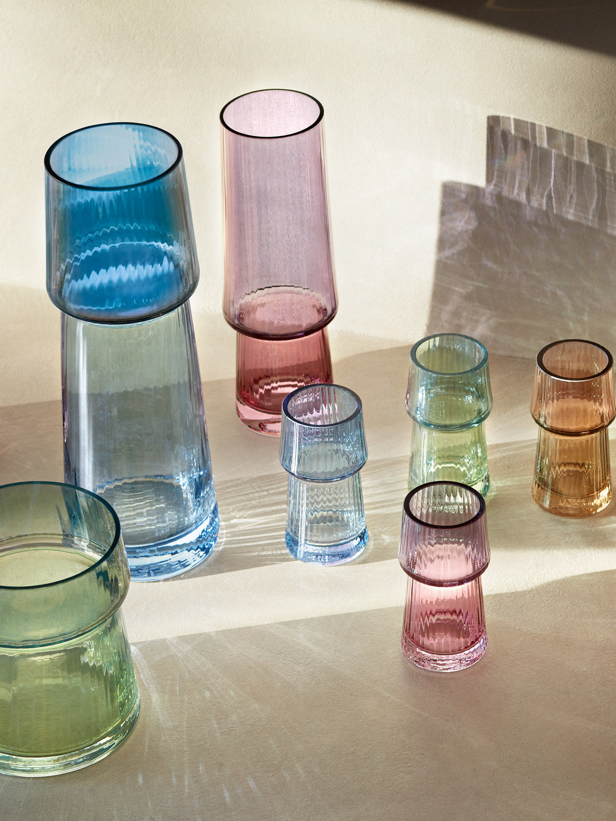 A selection of vibrant, mouth-blown glass vases by lsa-international, some displayed upside down, are arranged on a beige surface with sunlight casting colorful iridescent reflections and shadows around them.