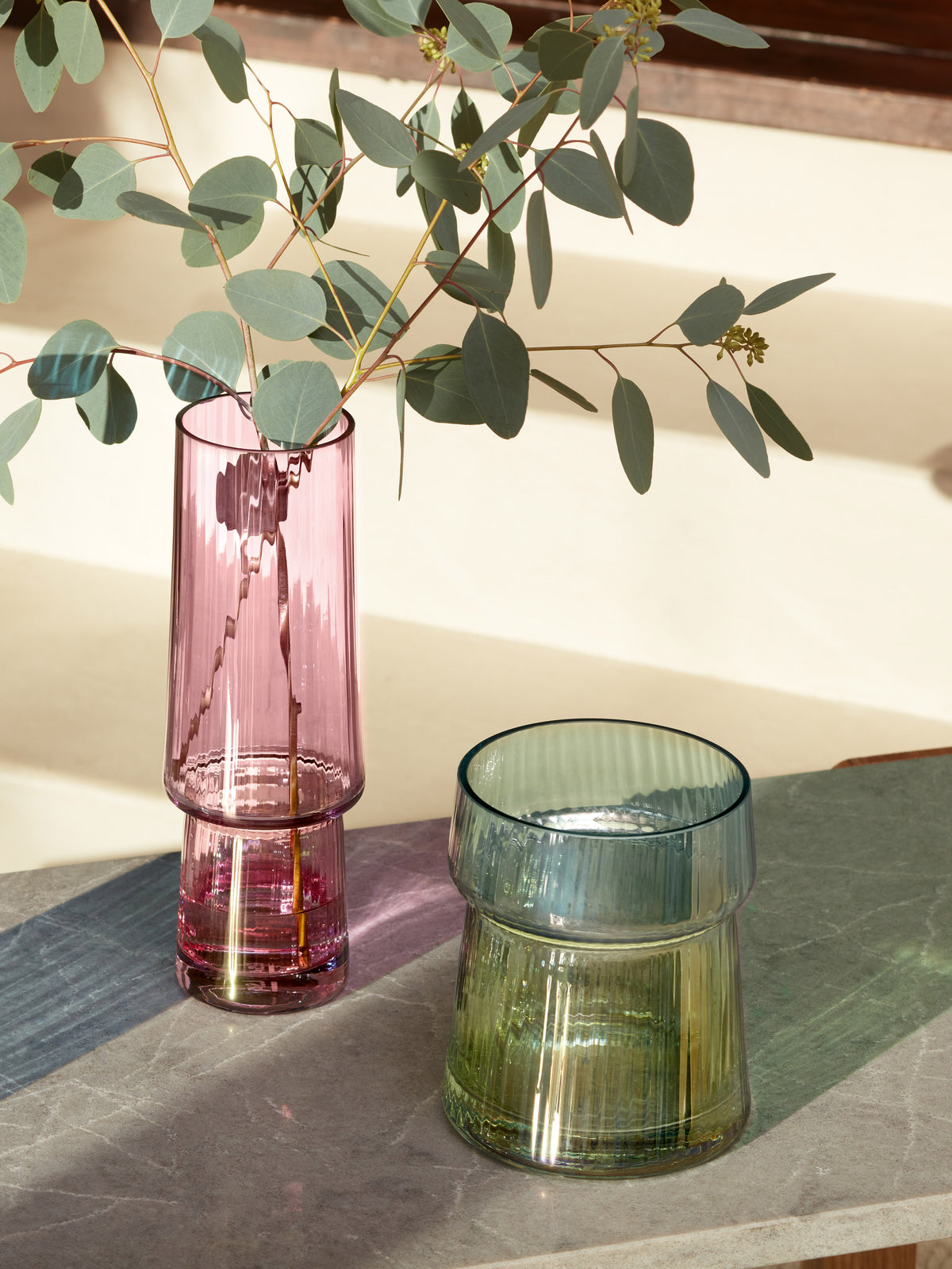 Two textured glass vases by lsa-international rest on a stone surface; one is a tall, iridescent pink lsa-international Vase filled with eucalyptus stems, while the other is a short green Vase from lsa-international. Sunlight casts soft shadows across the scene.