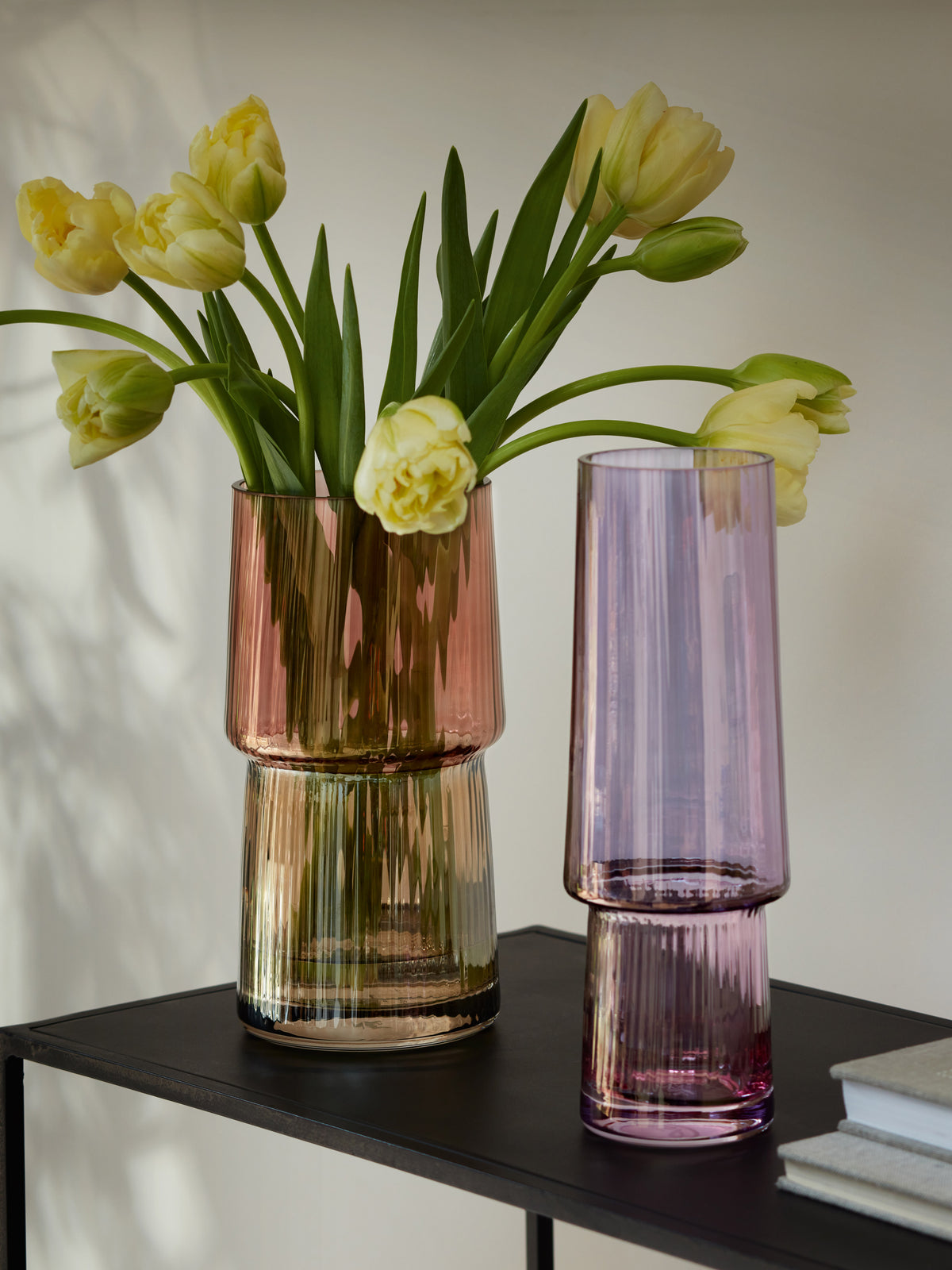 Two ribbed glass lsa-international Vases, one pink filled with yellow tulips and one purple, rest on a black metal table against a neutral wall. Each mouth-blown Vase from lsa-international catches the light beautifully. A stack of white books is partially visible to the right.