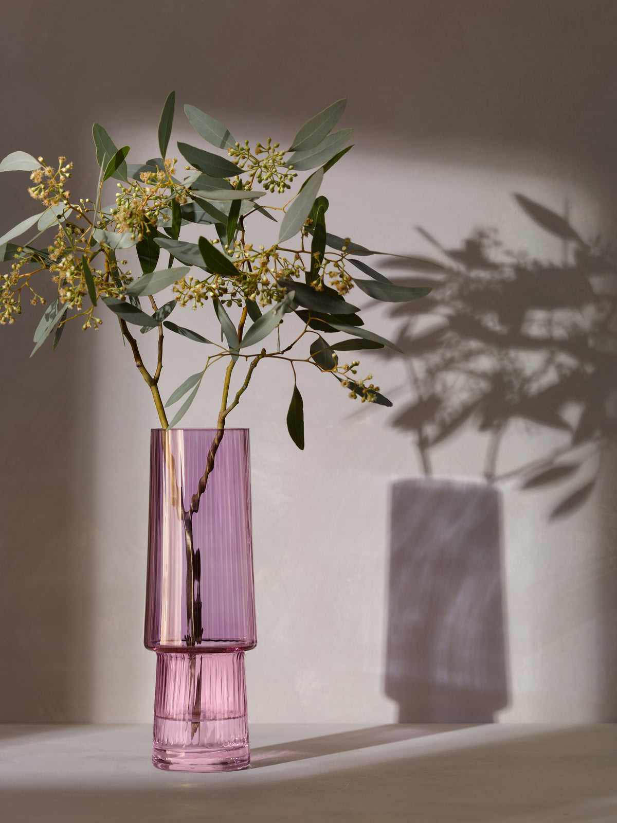 A mouth-blown pink Vase by lsa-international, filled with green leafy stems and small yellow flowers, casts a soft shadow on a light gray background.