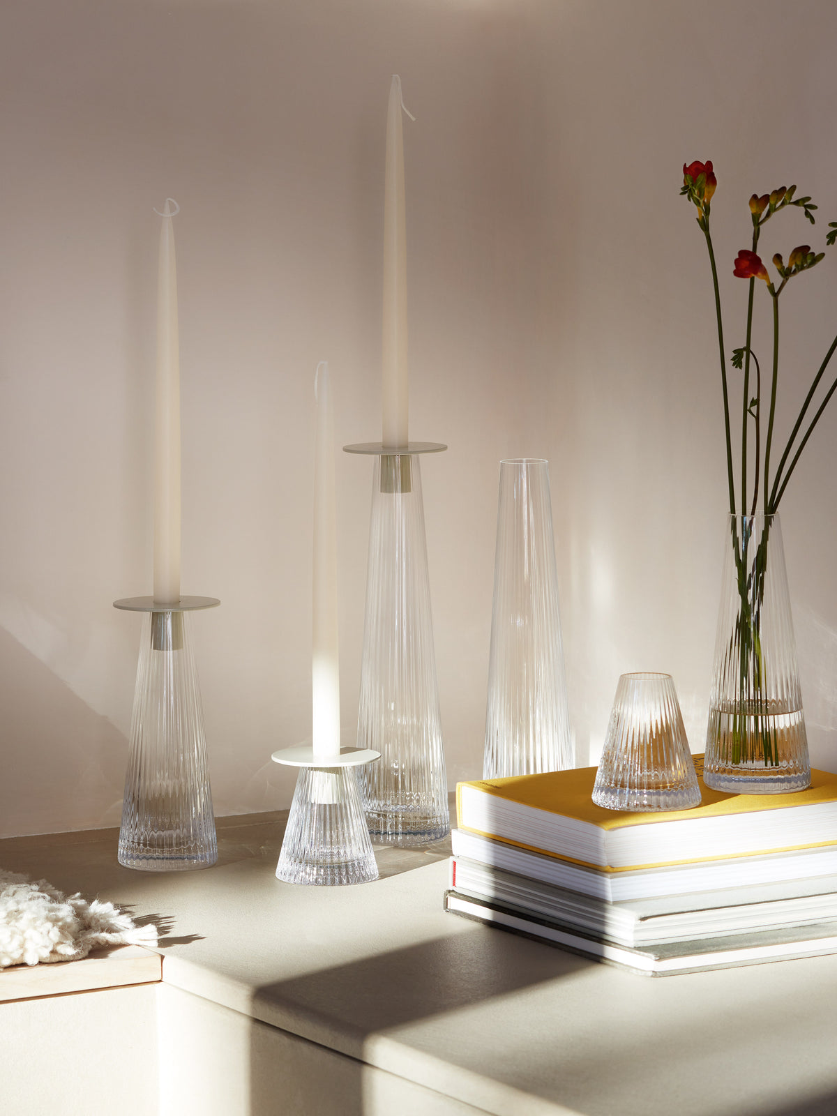Three LSA International Candleholder/Vase mouth-blown glass candleholders with tall taper candles and two pleated vases, one containing flowers, arranged on a stack of books.