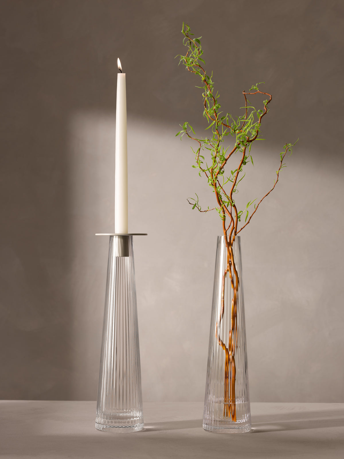 Two tall, pleated lsa-international Candleholder/Vase vessels: one holding a lit white taper candle and the other holding a branch with green leaves.