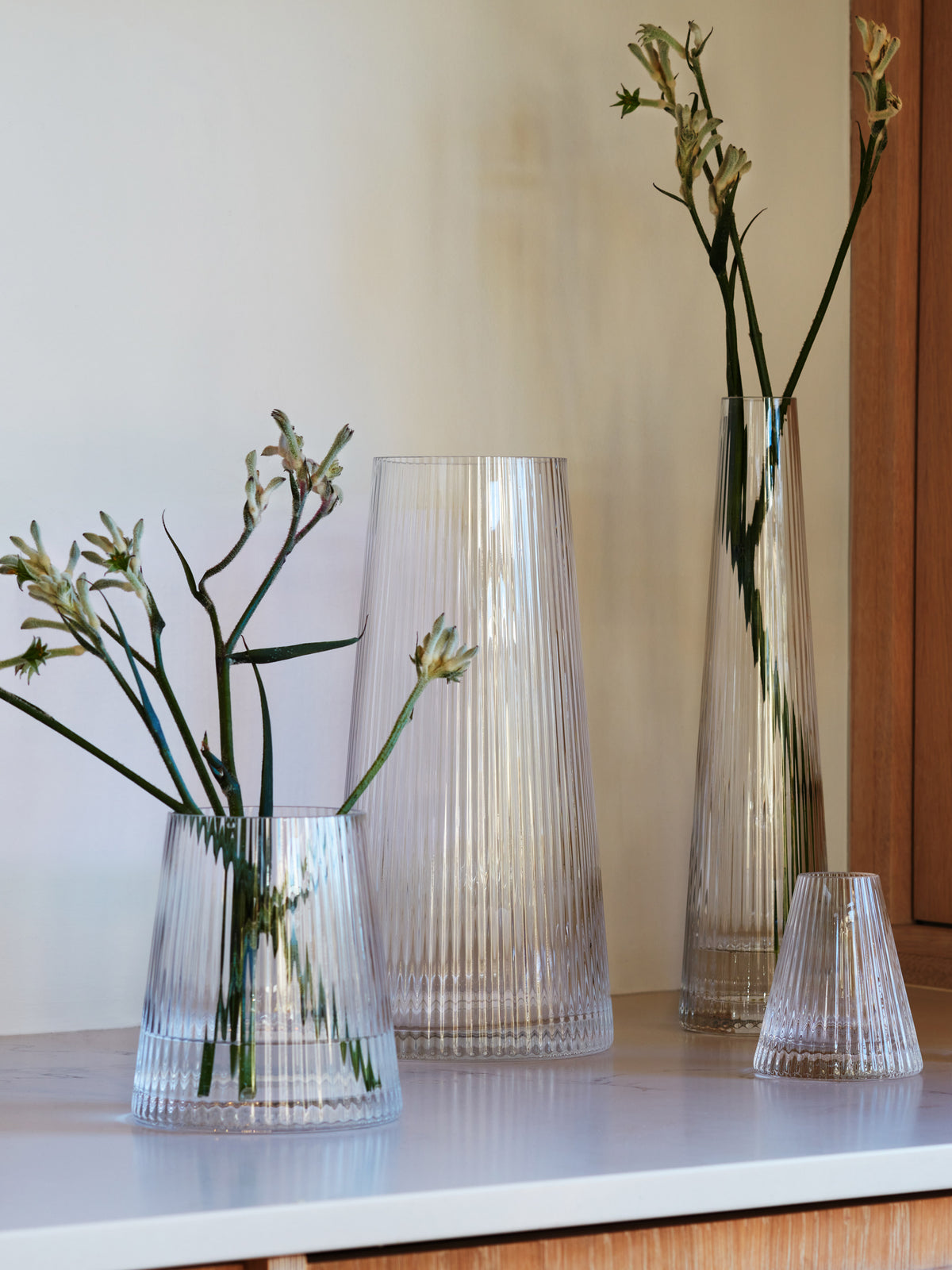Four LSA International Lantern/Vase vessels of varying heights arranged on a surface, including two pleated vases holding sprigs of green flowers.