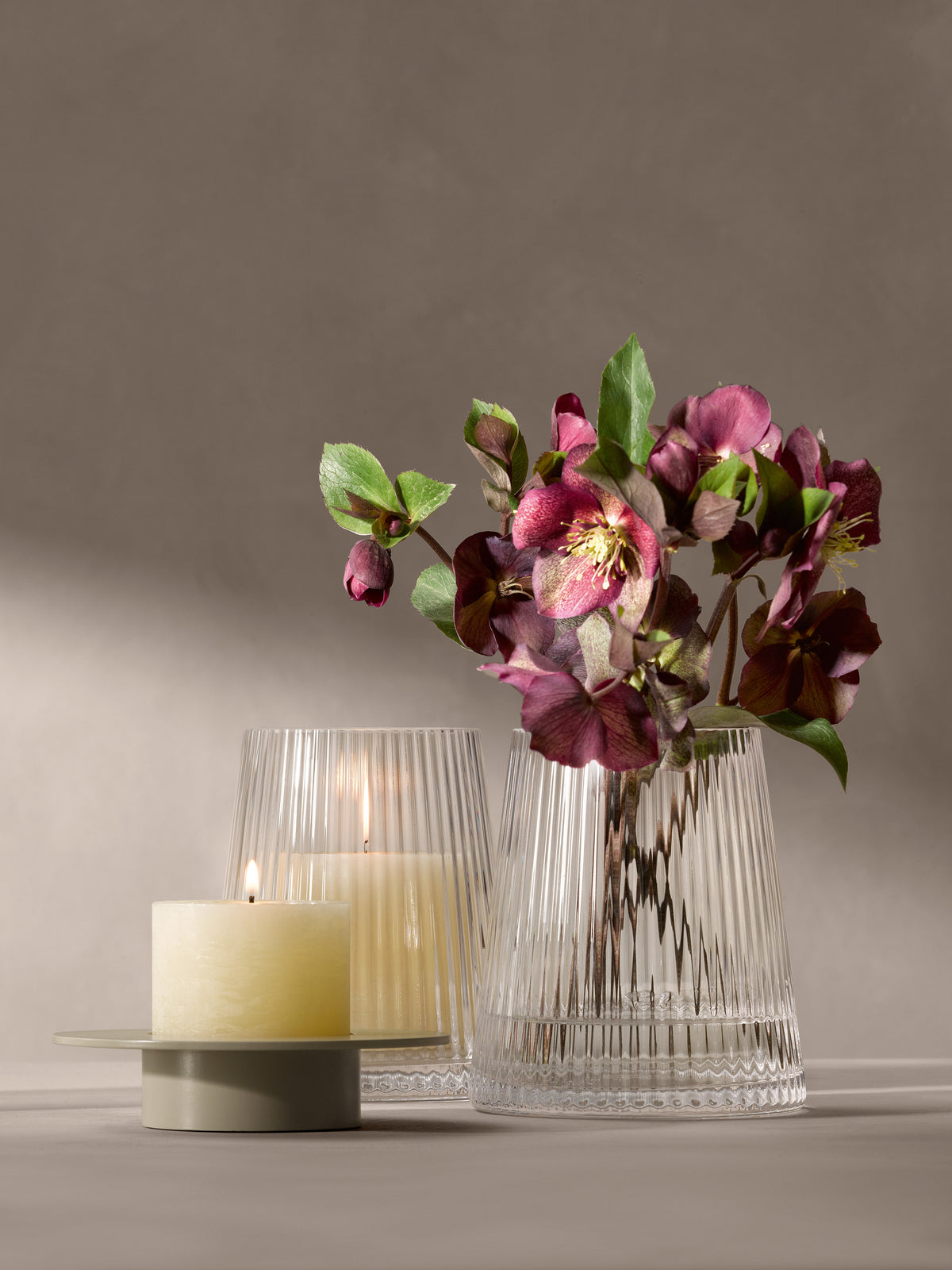 Two pleated glass Lantern/Vase pieces, one containing hellebore flowers and the other holding a lit pillar candle, next to a smaller candle on a low stand.
