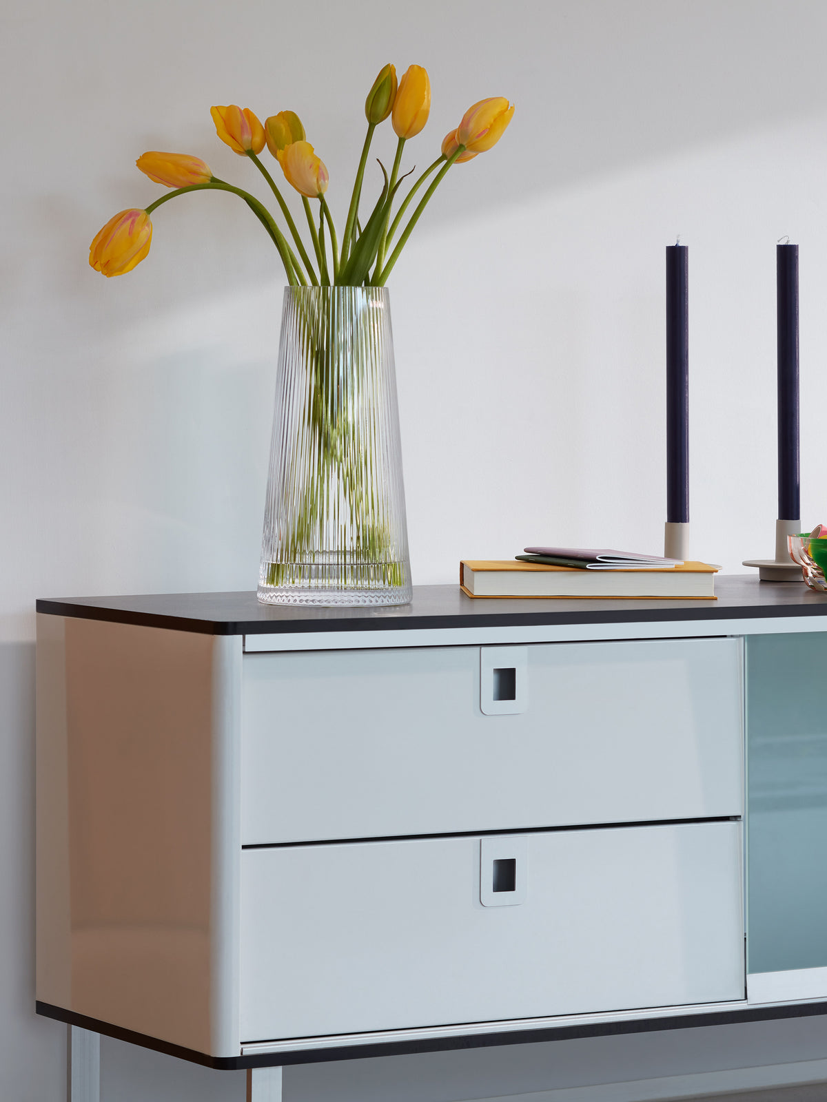 Clear, fluted LSA-International Lantern/Vase filled with yellow tulips sitting on a sideboard next to two navy blue taper candles in a modular candleholder and a stack of books, all illuminated by soft ambient light.