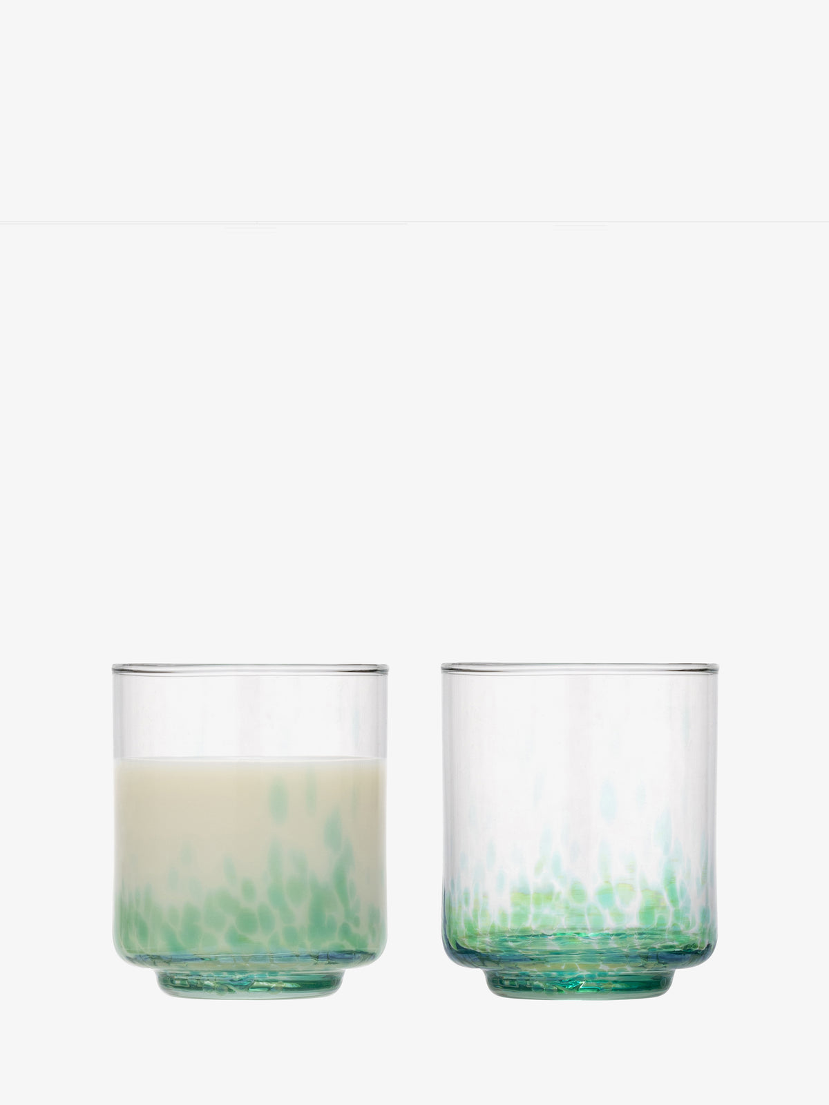 Two lsa-international Tumbler glasses made from clear recycled glass, each featuring a green speckled base, are displayed against a plain white background; the left tumbler is half-filled with milk, while the right one is empty.