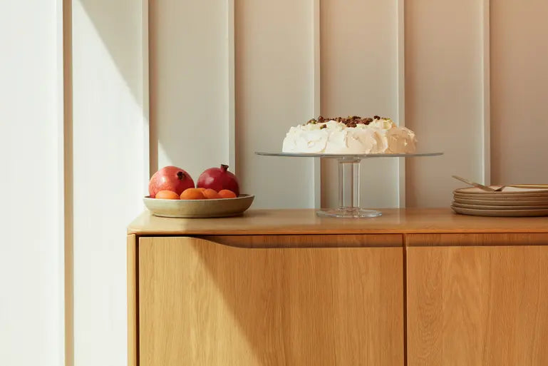 A wooden sideboard holds a plate of fruit, a cake on a glass stand, and a stack of plates, with sunlight casting shadows on a paneled white wall behind.