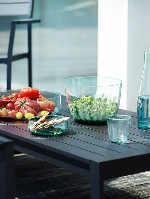 A dark outdoor table with a glass salad bowl, a bowl of assorted tomatoes, a plate of crackers, a glass, and a carafe of water. A chair and part of a sunlit patio are in the background.