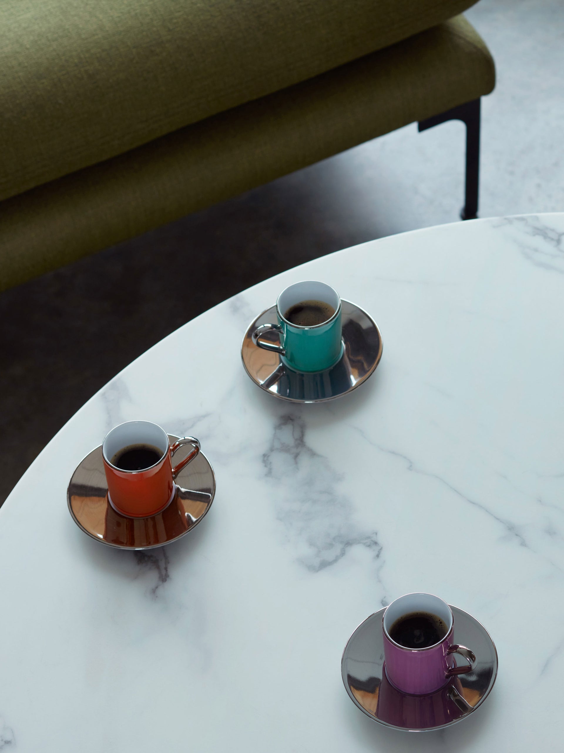 Three lsa-international Coffee Cup & Saucer sets—featuring iridescent, teal, and purple designs—are arranged on a round white marble table, with a green upholstered bench partially visible in the background.