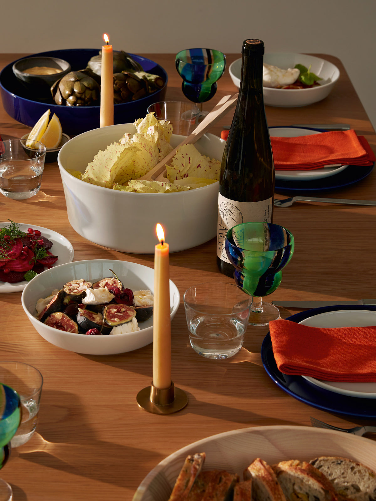 A large white LSA-International Pasta/Serving Bowl filled with radicchio salad sits on a wooden dining table set for a meal with plates, wine, candles, and various dishes.