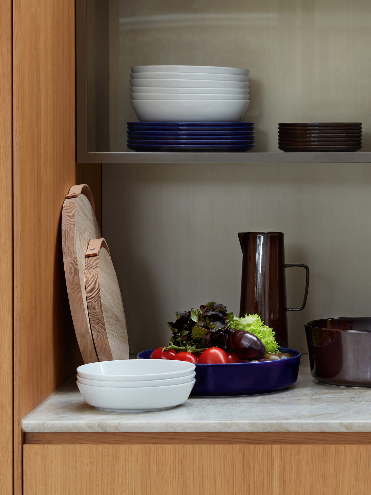 A blue LSA-International Pasta/Serving Bowl filled with salad greens and tomatoes sits on a kitchen counter next to white porcelain bowls, wooden serving boards, and a brown pitcher.