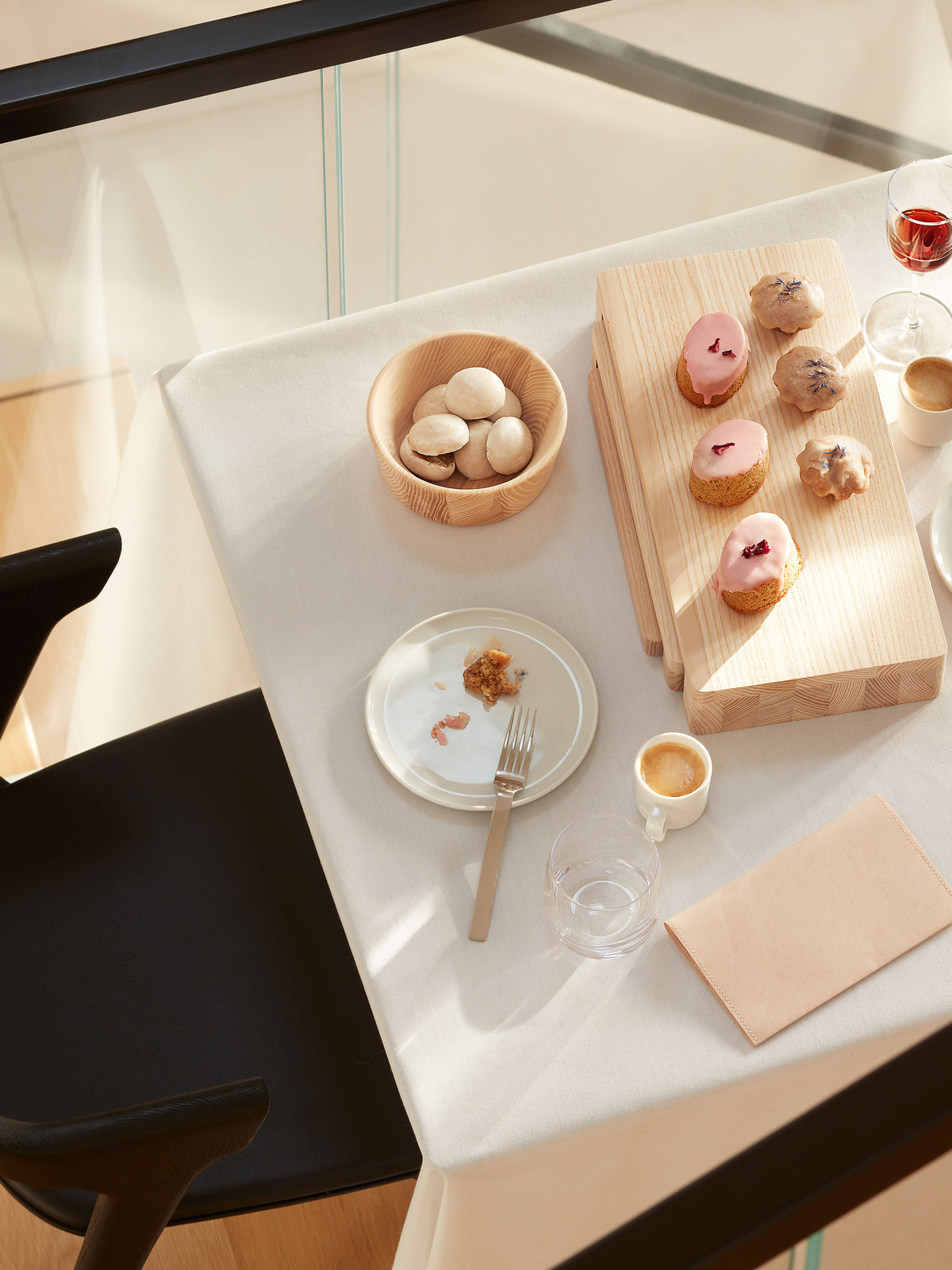 An LSA International Ash Wood Bowl filled with cookies and a contemporary serving board topped with glazed cakes set on a dining table with a plate, espresso, and glasses.