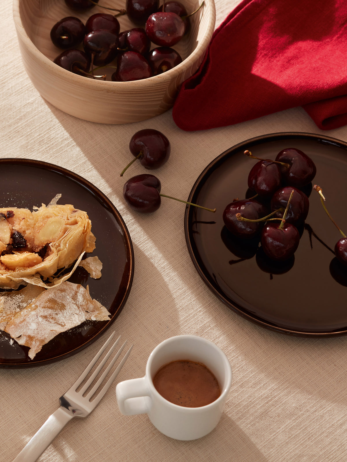 An LSA-International Ash Wood Bowl filled with cherries sits next to plates holding sliced strudel and fresh cherries, with a small cup of coffee and a fork nearby on a linen tablecloth.