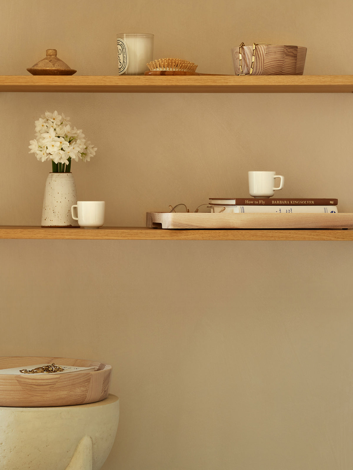 Two wooden shelves against a tan wall holding a white vase of flowers, books, small cups, and an lsa-international Ash Wood Bowl.