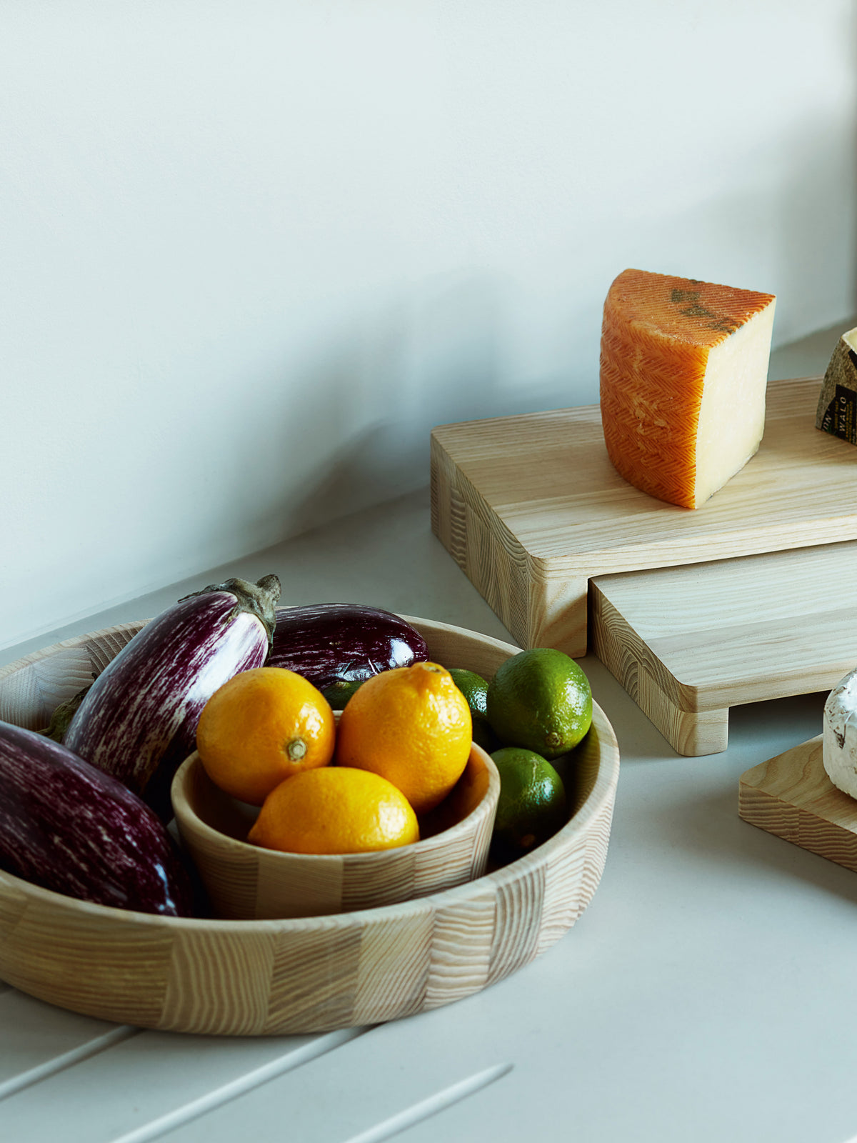 An LSA International Ash Wood Bowl filled with eggplants, lemons, and limes sits next to wooden serving boards topped with wedges of cheese.