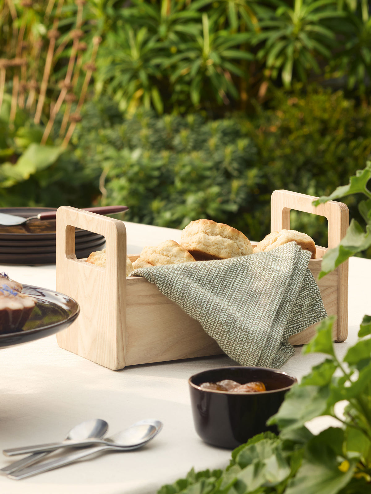 LSA International Ash Bread Basket, a rectangular untreated wood basket containing biscuits covered with a green patterned cloth, set on a table outdoors.