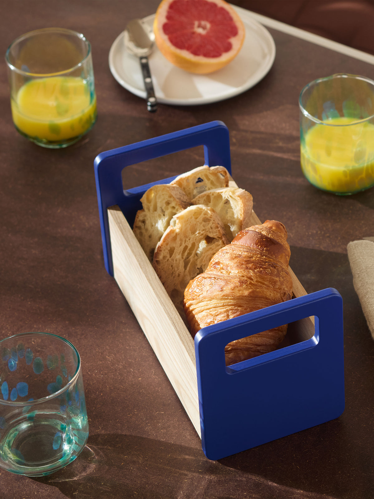 An lsa-international Ash Bread Basket with bright blue handles contains slices of bread and a croissant, set on a table with glasses of juice and a halved grapefruit.