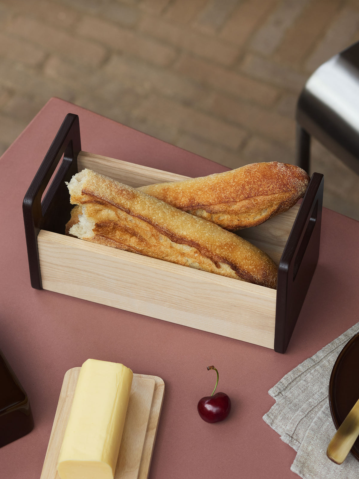 Two bread baguettes in an lsa-international Ash Bread Basket on a table next to a stick of butter and a single cherry.