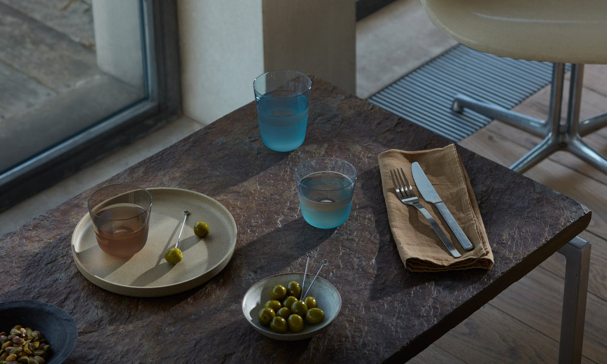 A stone table set with drinks served in the lsa-international Colour Tumbler Bundle - Low (6 piece) coloured glassware, accompanied by plates of green olives and nuts, a brown napkin with cutlery, positioned next to a window and a beige chair in a modern dining setting.