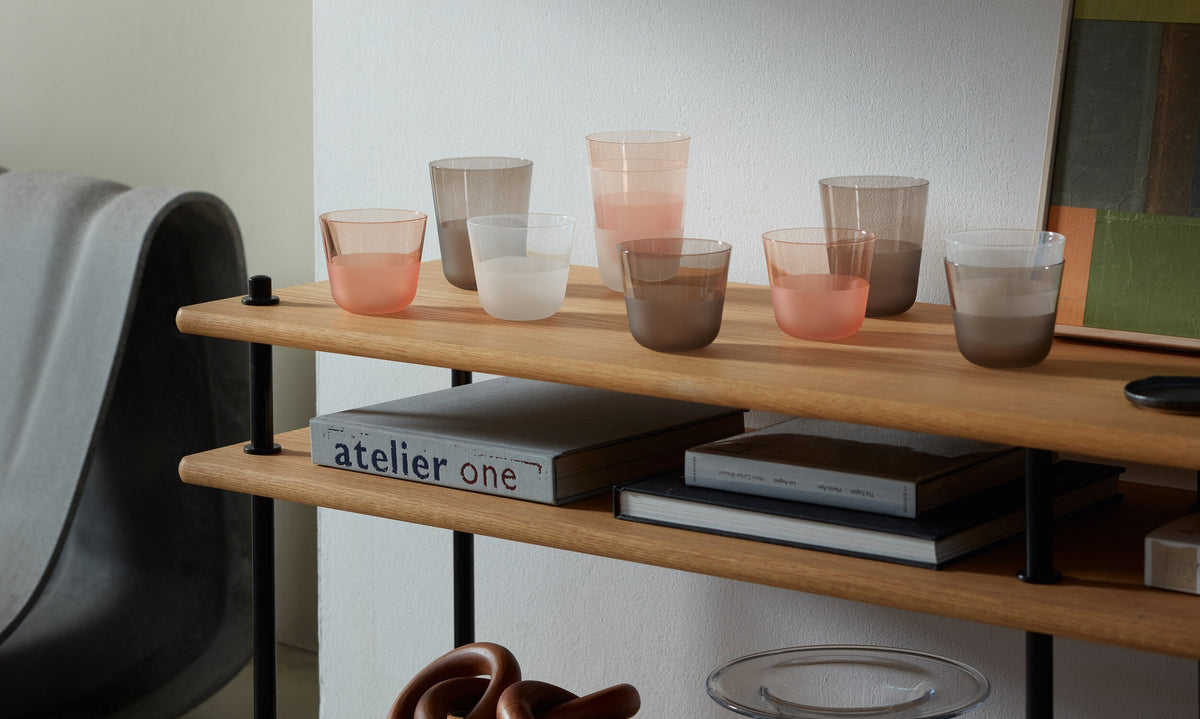 Six pastel-hued low tumblers from the lsa-international Colour Tumbler Bundle are arranged on a wooden shelf beside art books. The modern, minimalistic setting, with soft lighting and a neutral palette, accentuates the elegant design and stylish colored glassware.