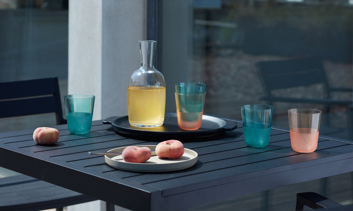 A black patio table from the Arc Contrast collection displays the lsa-international Colour Tumbler Bundle - Tall (6 Piece), arranged alongside a carafe of yellow beverage and three peaches on a plate. Sunlight illuminates the modern outdoor setup, emphasizing the matte textured finish of the table, with matching chairs and a building visible in the background.