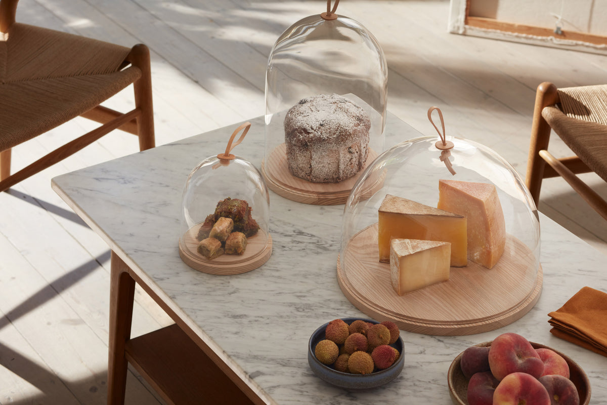 A marble table with three wooden chairs, covered with glass domes displaying cheeses, cake, pastries, and a bowl of lychees and peaches. Sunlight streams in through a window.