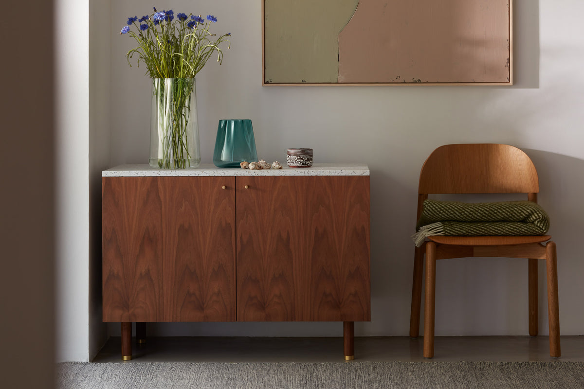 A wooden cabinet with a vase of flowers, a teal glass, and small decor on top stands next to a wooden chair with a folded green blanket, all against a light wall with a modern painting above.