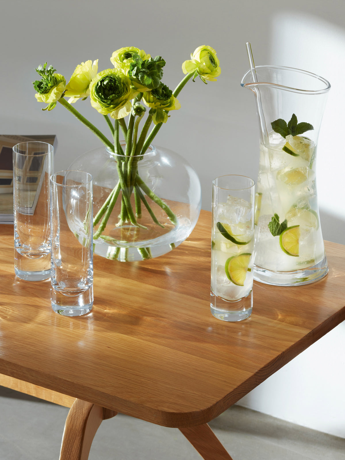 A wooden table holds a vase of yellow flowers, three empty Long Drink Glasses by lsa-international, and a glass pitcher of lemonade with lime, mint, and ice, all set in bright sunlight.