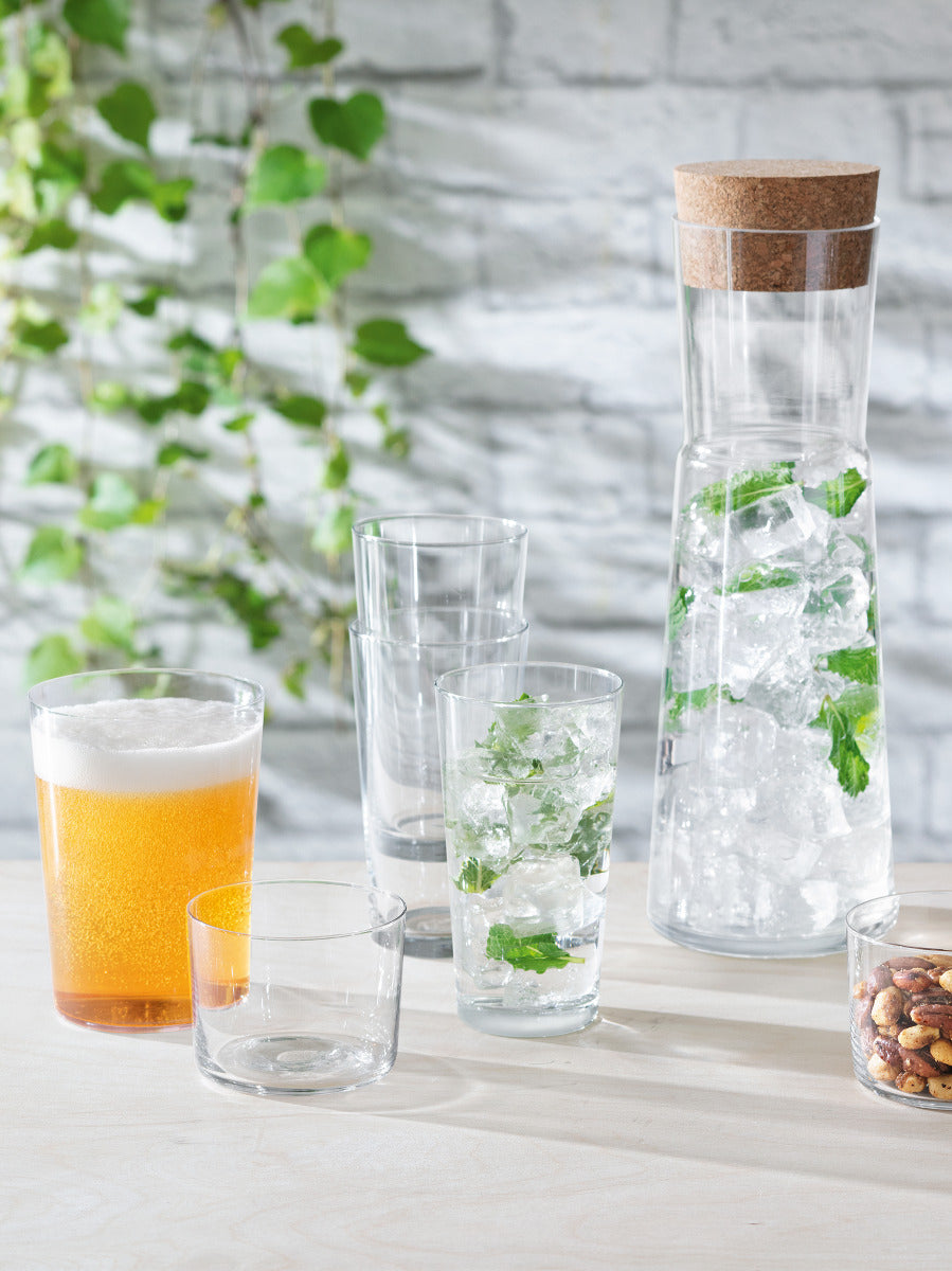 The lsa-international Carafe & Cork Stopper, filled with infused water, ice, and mint, sits on a table alongside three empty glasses, a glass of beer, a small bowl, and a bowl of mixed nuts. Green leaves and a white brick wall provide the backdrop.