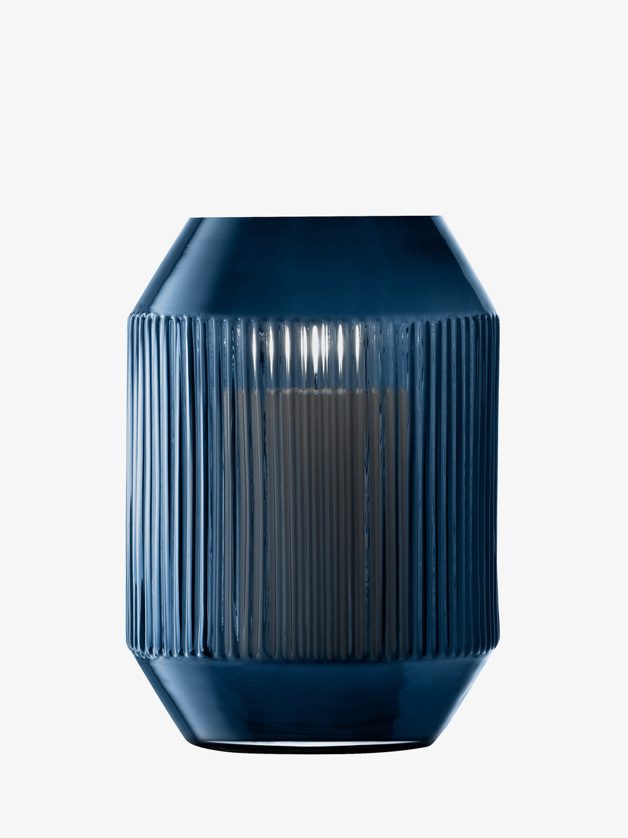 The lsa-international Lantern/Vase features a blue ribbed ceramic design with a glossy finish and contains a lit white pillar candle inside. Showcasing a modern, geometric silhouette inspired by mouth-blown sapphire blue glass, this piece is presented against a plain white background.