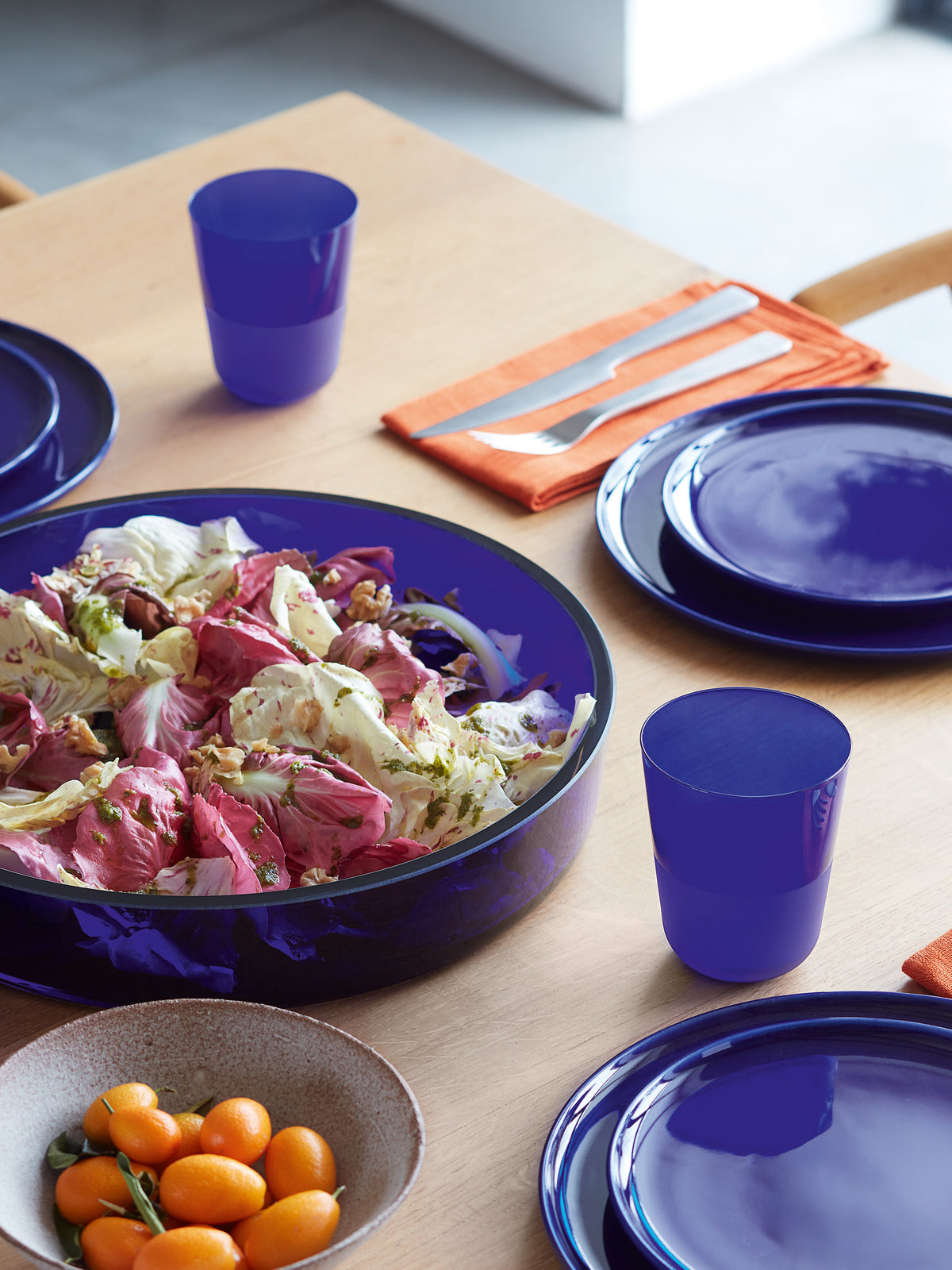 A table set for four with blue plates and cups featuring a matte textured finish, an orange napkin with cutlery, a salad in a large blue bowl, lsa-international Tumbler soda-lime glass tumblers, and a small bowl of oranges.