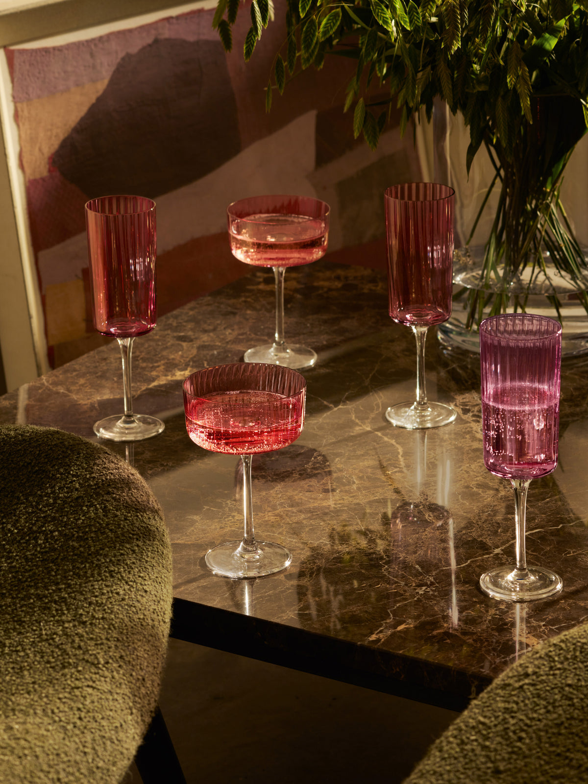 Five pink and purple ribbed glass goblets and coupes sit on a dark marble table, accompanied by lsa-international Champagne Flutes that catch the light, with green chairs and a vase of greenery in the background. Sunlight casts warm shadows across the scene.